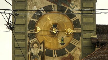 Eine große, historische Uhr ist an der Wand eines Gebäudes zu sehen. Der Ziffernblatt zeigt goldene Zeiger und ist reich verziert mit Bildern von einem Engel und einer Figur. Die Uhr ist in einem städtischen Umfeld mit Stromleitungen im Vordergrund platziert.