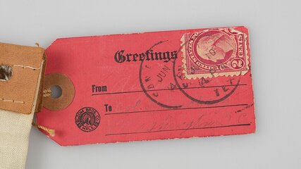 Ein rotes Etikett mit der Aufschrift "Greetings". Oben ist ein Poststempel zu sehen, der den Monat Juni markiert. Es gibt Bereiche für "Von" und "An", die handschriftlich ausgefüllt sind. Das Etikett hat eine braune Lederbefestigung.