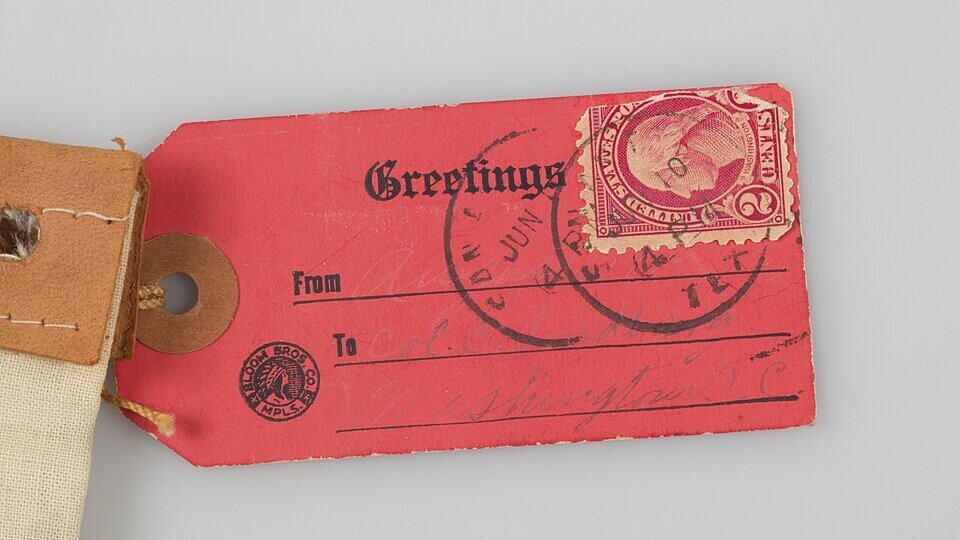 Ein rotes Etikett mit der Aufschrift "Greetings". Oben ist ein Poststempel zu sehen, der den Monat Juni markiert. Es gibt Bereiche für "Von" und "An", die handschriftlich ausgefüllt sind. Das Etikett hat eine braune Lederbefestigung.