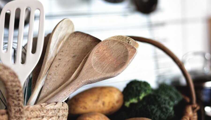 Eine Korb mit verschiedenen Kochlöffeln, darunter Holzlöffel und ein Pfannenwender, steht neben frischem Gemüse, darunter Brokkoli und Kartoffeln. Die Utensilien deuten auf das Zubereiten von Mahlzeiten hin.