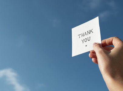 Eine Hand hält ein kleines Blatt Papier mit der Aufschrift „THANK YOU“. Der Hintergrund zeigt einen klaren blauen Himmel.