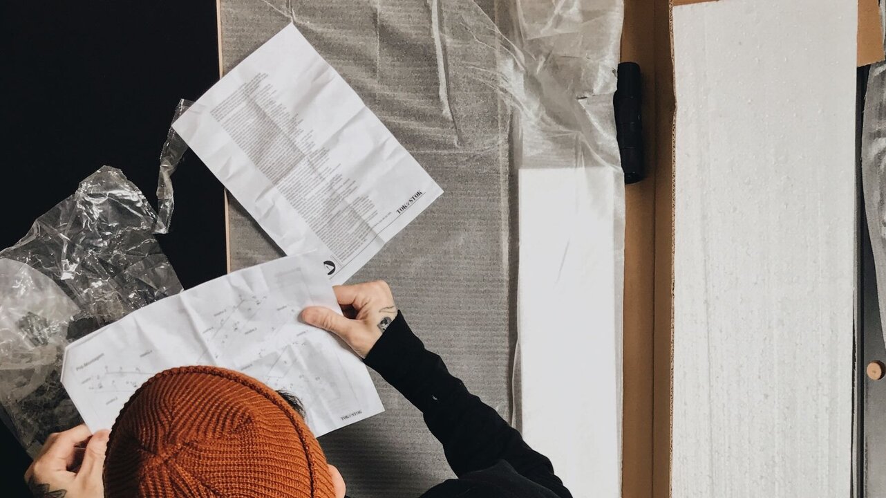Eine Person mit einem braunen Beanie sitzt über einem Karton und hält Anleitungen in der Hand. Um sie herum befinden sich Verpackungsmaterialien und Teile. Der Fokus liegt auf dem Lesen der Anleitung, während der Raum unordentlich aussieht.