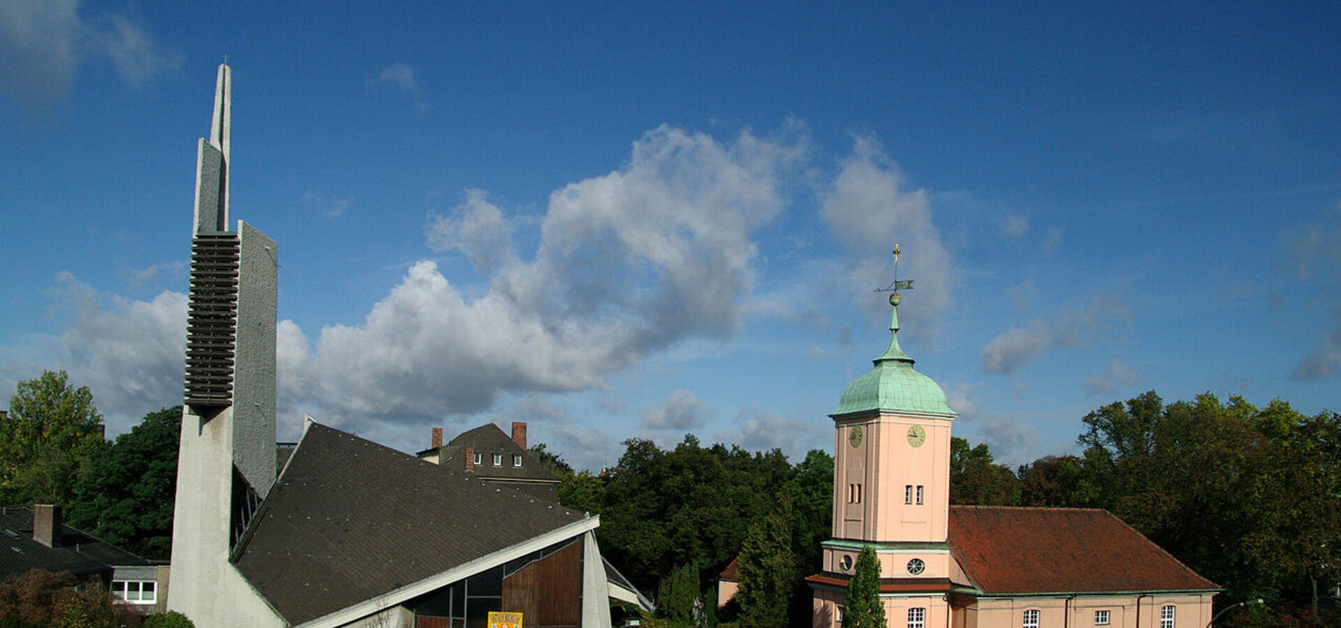 Bildbeschreibung: Zwei Kirchengebäude in einer grünen Umgebung. Links ein modernes, spitzes Bauwerk mit einer hohen Turmhaube, rechts eine klassische, rosa Kirche mit grüner Kuppel. Der Himmel ist blau mit einigen Wolken.