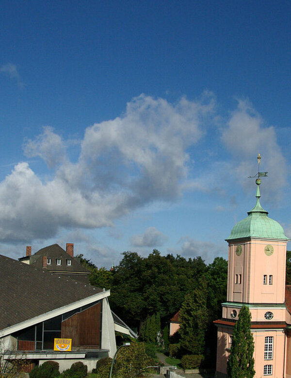 Bildbeschreibung: Zwei Kirchengebäude in einer grünen Umgebung. Links ein modernes, spitzes Bauwerk mit einer hohen Turmhaube, rechts eine klassische, rosa Kirche mit grüner Kuppel. Der Himmel ist blau mit einigen Wolken.