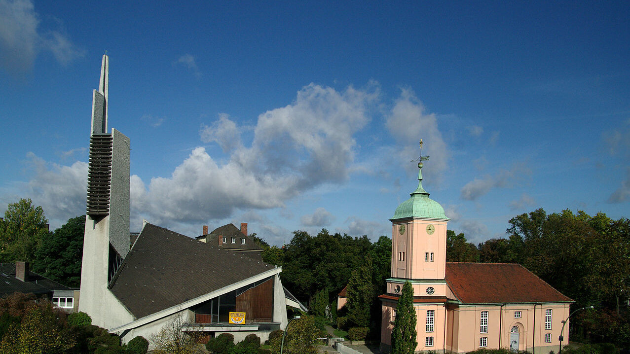 Bildbeschreibung: Zwei Kirchengebäude in einer grünen Umgebung. Links ein modernes, spitzes Bauwerk mit einer hohen Turmhaube, rechts eine klassische, rosa Kirche mit grüner Kuppel. Der Himmel ist blau mit einigen Wolken.