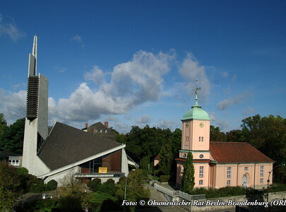 Bildbeschreibung: Zwei Kirchengebäude in einer grünen Umgebung. Links ein modernes, spitzes Bauwerk mit einer hohen Turmhaube, rechts eine klassische, rosa Kirche mit grüner Kuppel. Der Himmel ist blau mit einigen Wolken.