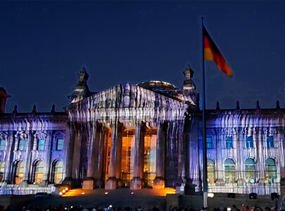 Das Bild zeigt das Reichstagsgebäude in Berlin, beleuchtet durch eine farbige Projektion, während die Dämmerung einsetzt. Vor dem Gebäude sind Menschen versammelt, die die Lichtshow bewundern. Auf den Dächern wehen deutsche Flaggen.