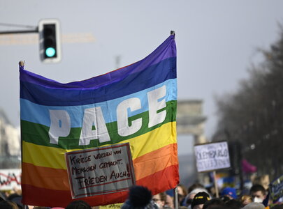 Ein Regenbogen-Flagge mit der Aufschrift "PACE" steht im Vordergrund einer Menschenmenge. Darunter ein Schild mit der Botschaft: "Kriege werden von Menschen gemacht. Frieden auch!" Im Hintergrund sind verschwommen Menschen und eine Landmarke zu sehen.