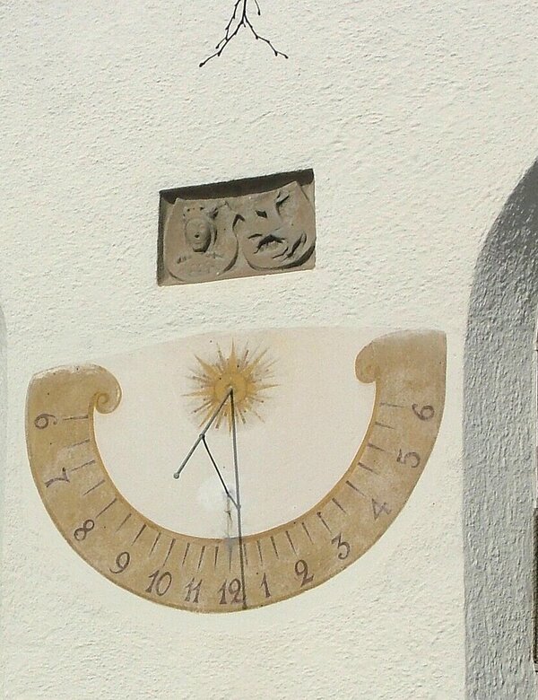Ein Sonnenstundenmesser ist an einer weißen Wand mit zwei Fenstern angebracht. Der Stundenmesser zeigt die Ziffern von 1 bis 12, mit einem Sonnenmotif in der Mitte und dem Zeiger, der die aktuelle Zeit anzeigt.