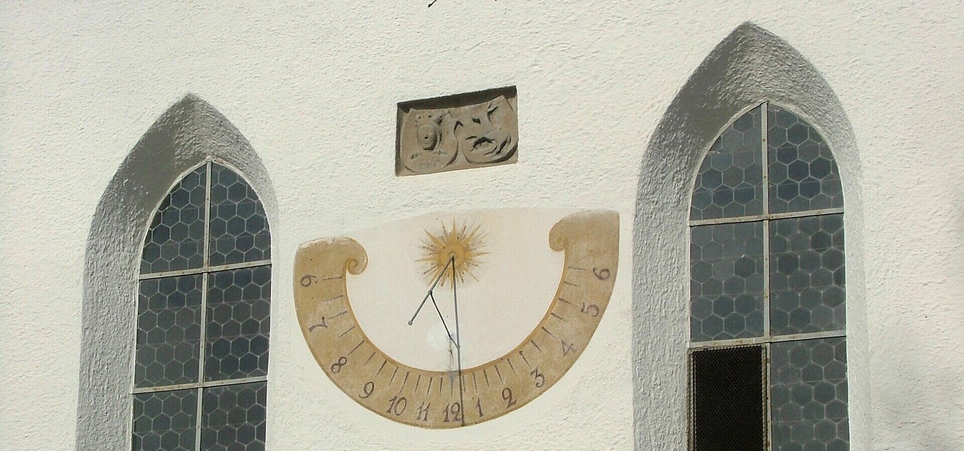 Ein Sonnenstundenmesser ist an einer weißen Wand mit zwei Fenstern angebracht. Der Stundenmesser zeigt die Ziffern von 1 bis 12, mit einem Sonnenmotif in der Mitte und dem Zeiger, der die aktuelle Zeit anzeigt.
