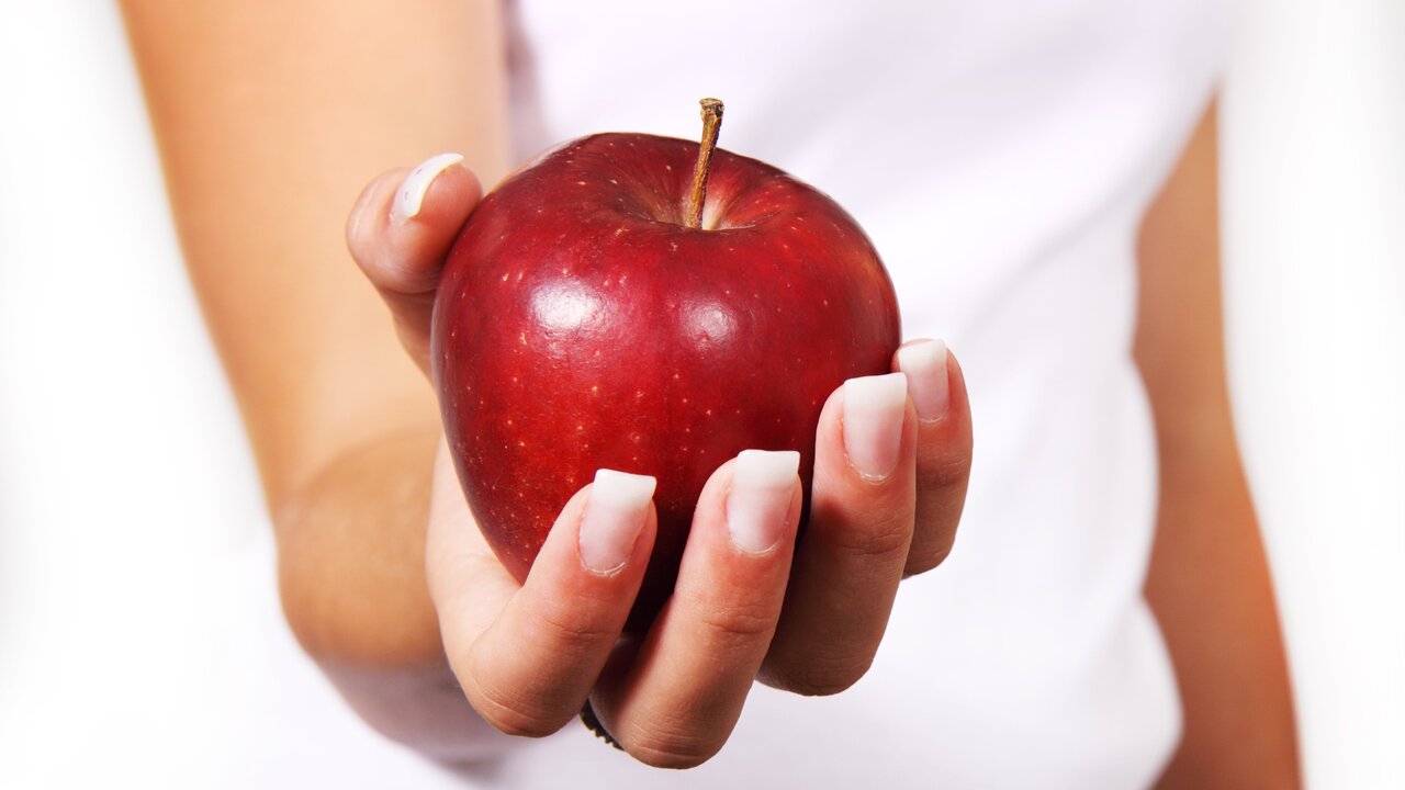 Eine Hand hält einen glänzenden, roten Apfel. Der Hintergrund ist hell und neutral, wodurch der Apfel im Mittelpunkt steht.