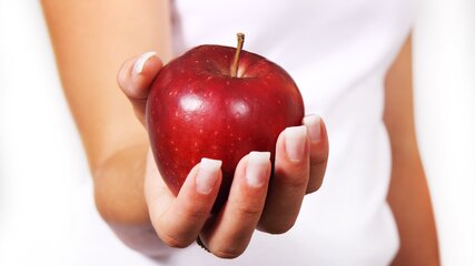 Eine Hand hält einen glänzenden, roten Apfel. Der Hintergrund ist hell und neutral, wodurch der Apfel im Mittelpunkt steht.