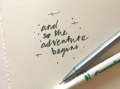 Ein offenes Notizbuch liegt auf einem Tisch. Darin steht handschriftlich der Satz: „and so the adventure begins“. Neben dem Buch sind ein grüner und ein silberner Stift platziert, die den kreativen Schreibprozess symbolisieren.