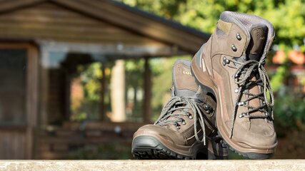 Zwei Wanderstiefel stehen auf einer Holzoberfläche. Die Stiefel sind braun und verfügen über starke Schnürsenkel sowie einen hohen Schaft, der Knöchelschutz bietet. Im Hintergrund ist eine unscharfe Holzstruktur zu erkennen, die auf eine Hütte oder ein Gebäude hindeutet.