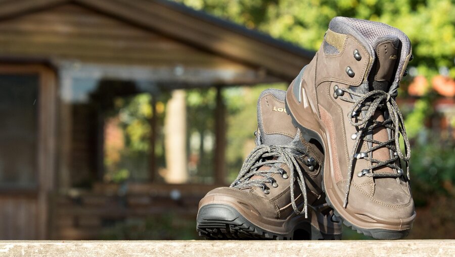 Zwei Wanderstiefel stehen auf einer Holzoberfläche. Die Stiefel sind braun und verfügen über starke Schnürsenkel sowie einen hohen Schaft, der Knöchelschutz bietet. Im Hintergrund ist eine unscharfe Holzstruktur zu erkennen, die auf eine Hütte oder ein Gebäude hindeutet.
