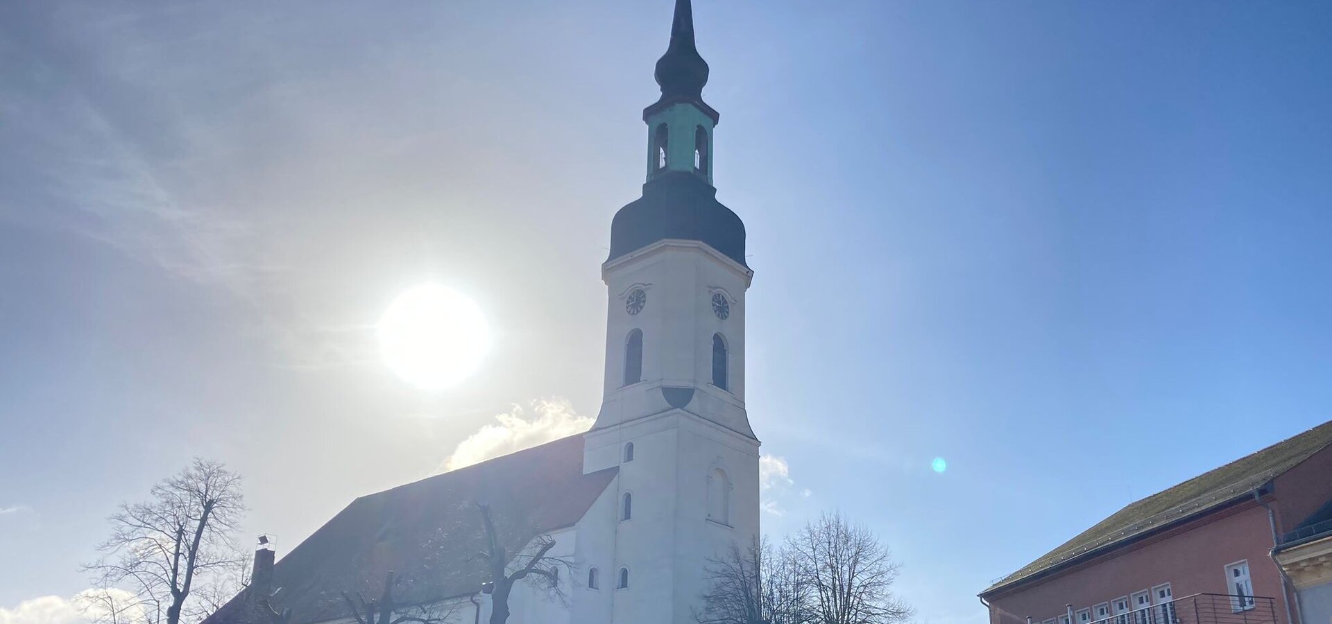 Eine weiße Kirche mit einem hohen, spitzen Turm steht im Vordergrund. Die Sonne scheint hell im Hintergrund, und der Himmel ist klar mit einigen Wolken. Um die Kirche sind einige Bäume sichtbar, und es gibt Parkplätze in der Nähe.