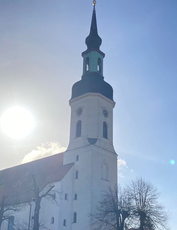 Eine weiße Kirche mit einem hohen, spitzen Turm steht im Vordergrund. Die Sonne scheint hell im Hintergrund, und der Himmel ist klar mit einigen Wolken. Um die Kirche sind einige Bäume sichtbar, und es gibt Parkplätze in der Nähe.