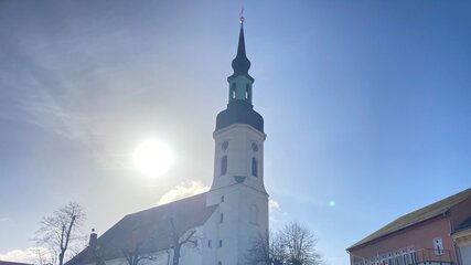 Eine weiße Kirche mit einem hohen, spitzen Turm steht im Vordergrund. Die Sonne scheint hell im Hintergrund, und der Himmel ist klar mit einigen Wolken. Um die Kirche sind einige Bäume sichtbar, und es gibt Parkplätze in der Nähe.