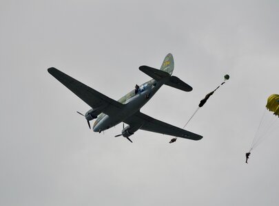 Ein Flugzeug schwebt über dem Himmel, während mehrere Fallschirmspringer aussteigen. Einer der Springer hat einen gelben Fallschirm geöffnet und gleitet nach unten, während ein weiterer in der Luft hängt, bereit zum Sprung. Die Wolken sind grau und vermitteln eine dynamische, aufregende Atmosphäre.