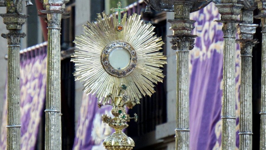 Ein goldener und silberner Monstranz mit strahlenden Sonnenstrahlen, die eine kleine Hostie in der Mitte halten. Im Hintergrund sind dekorative purpurne Stoffe sichtbar. Die Monstranz wird oft in religiösen Zeremonien verwendet, um die Eucharistie zu präsentieren.