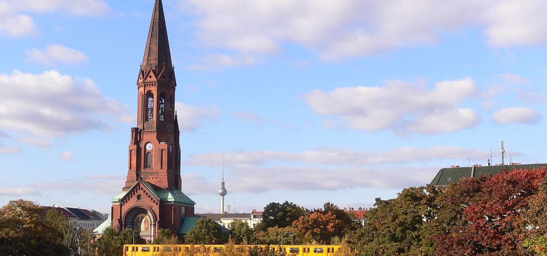 Eine rote Backsteinkirche mit spitzem Turm steht im Vordergrund, während im Hintergrund ein gelber Zug vorbeifährt. Bunte Herbstbäume umrahmen die Szene, mit einem klaren Himmel im Hintergrund.