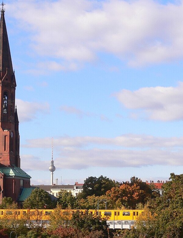 Eine rote Backsteinkirche mit spitzem Turm steht im Vordergrund, während im Hintergrund ein gelber Zug vorbeifährt. Bunte Herbstbäume umrahmen die Szene, mit einem klaren Himmel im Hintergrund.
