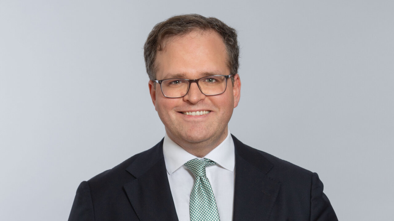 Ein lächelnder Mann in einem dunkelblauen Anzug und einer grünen Krawatte steht vor einem hellgrauen Hintergrund. Er trägt eine Brille und hat die Hände leicht vor sich. Seine Körpersprache strahlt Selbstbewusstsein und Freundlichkeit aus.