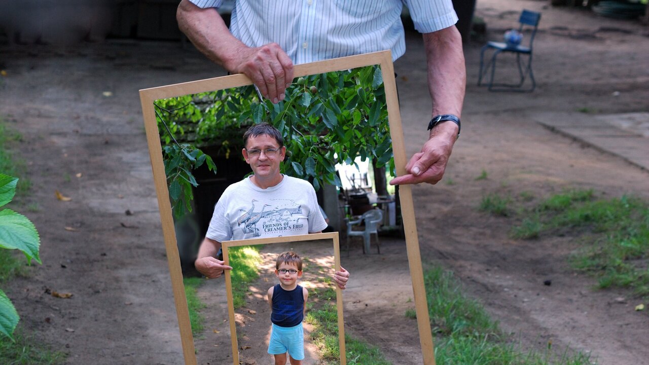 Ein älterer Mann steht im Freien und hält einen Bilderrahmen, in dem zwei Fotos zu sehen sind: eines von ihm als Kind und eines von ihm als Erwachsenem. Im Hintergrund sind Bäume und eine besondere ländliche Umgebung sichtbar.
