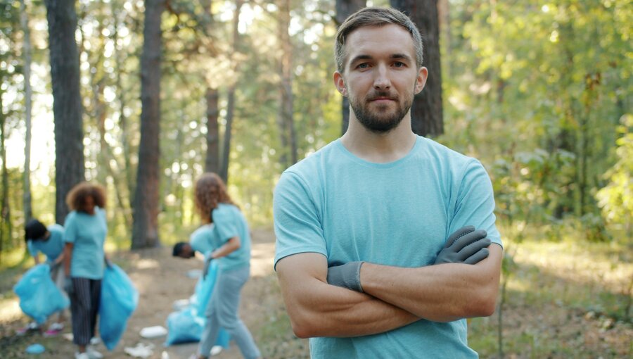 Eine Gruppe von Menschen führt Müllsammlung in einem Waldgebiet durch. Im Vordergrund steht ein junger Mann in einem hellblauen T-Shirt mit verschränkten Armen und einem entschlossenen Gesichtsausdruck. Hinter ihm sind zwei Personen, die Müllsäcke füllen.