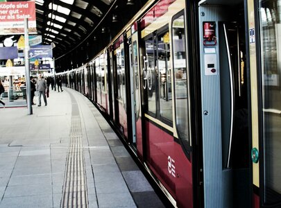 Ein Bahnhof mit einer einfahrenden Bahn, die im Vordergrund sichtbar ist. Passagiere stehen am Gleis und warten auf den Zug. Die Architektur des Bahnhofs ist modern mit einem wellenförmigen Dach. Hinweise und Anzeigetafeln sind im Hintergrund sichtbar.