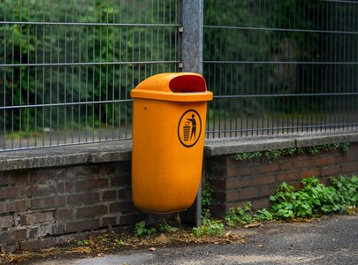 Ein orangefarbener Müllcontainer steht an einer Wand. Er hat einen Deckel mit einer Öffnung für Abfälle und ein Piktogramm, das das Entsorgen von Müll symbolisiert. Im Hintergrund sind Pflanzen und eine Zaunanlage sichtbar.