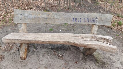 Eine verwitterte Holzbank steht in einer Waldumgebung. Auf der Rückenlehne ist der Schriftzug "FREE LUIT" sichtbar. Der Boden ist sandig und von herabgefallenem Laub umgeben.