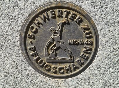Ein runder bronzefarbener Medaillon mit dem Text "SCHWERTER ZU PFLUGSCHAREN" und einem stilisierten, kämpfenden Menschen, der ein Schwert in der Hand hebt. Unten steht "MICA 4:3".