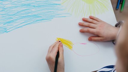 Ein Kind zeichnet auf einem großen Blatt Papier. Es ist mit verschiedenen Farben aktiv, darunter Blau für Wasser und Gelb für die Sonne. Die Hände des Kindes sind in Aktion, während es eine runde Form in Pink und eine gelbe Farbe nutzt.