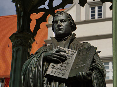 Eine Statue von Martin Luther, der ein Buch in der Hand hält. Sein Gesichtsausdruck ist ernst und nachdenklich. Die Statue steht in einem überdachten Bereich, umgeben von architektonischen Elementen. Luther trägt traditionelle Kleidung der Reformationszeit und wird oft mit seiner Rolle in der christlichen Reformbewegung assoziiert.
