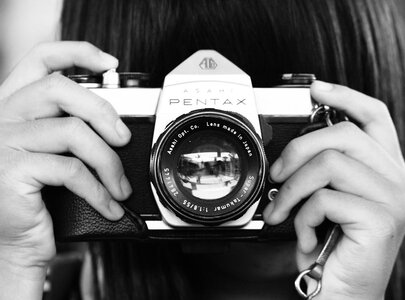 Eine Person hält eine Pentax-Kamera vor ihr Gesicht, bereit, ein Foto zu machen. Die Kamera hat ein klassisches Design mit einem runden Objektiv und ist in Schwarz-Weiß dargestellt.