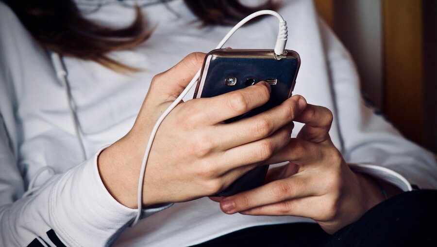 Eine Person hält ein Smartphone mit einem angeschlossenen Kopfhörer. Die Hände sind gut sichtbar, während das Smartphone zwischen ihnen gehalten wird. Die Person trägt ein weißes Sweatshirt mit einem schwarzen Schriftzug.