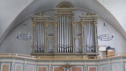 Eine prunkvolle Orgel in einem Kirchenraum. Die Orgel hat mehrere Pfeifenreihen und ist kunstvoll verziert. Der Hintergrund ist schlicht gehalten, mit einem hohen Gewölbe und hängenden Leuchten. Die Orgel steht auf einer erhöhten Plattform, was ihr einen markanten Platz im Raum verleiht.