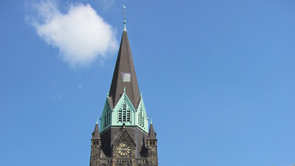 Eine alte Kirche mit einem hohen, spitzen Turm und grünen Dächern steht im Vordergrund. Die Straße davor ist von Bäumen gesäumt, und der Himmel ist klar und blau. Ein großes, rot gestaltetes Objekt ist am Ende der Straße sichtbar.