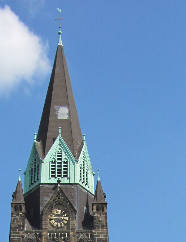 Eine alte Kirche mit einem hohen, spitzen Turm und grünen Dächern steht im Vordergrund. Die Straße davor ist von Bäumen gesäumt, und der Himmel ist klar und blau. Ein großes, rot gestaltetes Objekt ist am Ende der Straße sichtbar.