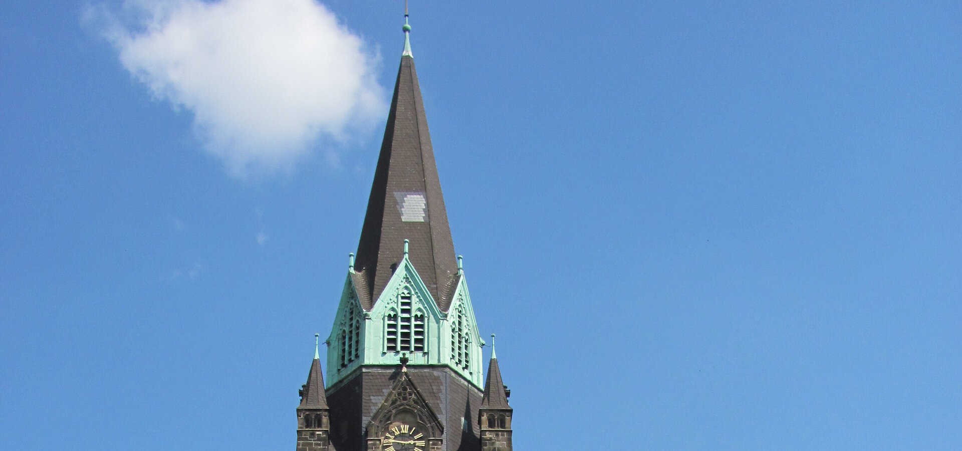 Eine alte Kirche mit einem hohen, spitzen Turm und grünen Dächern steht im Vordergrund. Die Straße davor ist von Bäumen gesäumt, und der Himmel ist klar und blau. Ein großes, rot gestaltetes Objekt ist am Ende der Straße sichtbar.