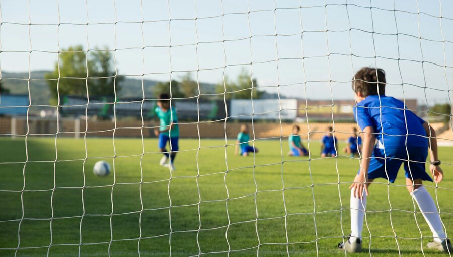 Eine Gruppe von Kindern spielt Fußball auf einem Rasenplatz. Im Vordergrund ist ein Junge in blauer Kleidung zu sehen, der dem Ball zuschaut. Im Hintergrund üben andere Kinder. Der Ball ist in Richtung Tor positioniert, das von einem Tornetz umgeben ist.