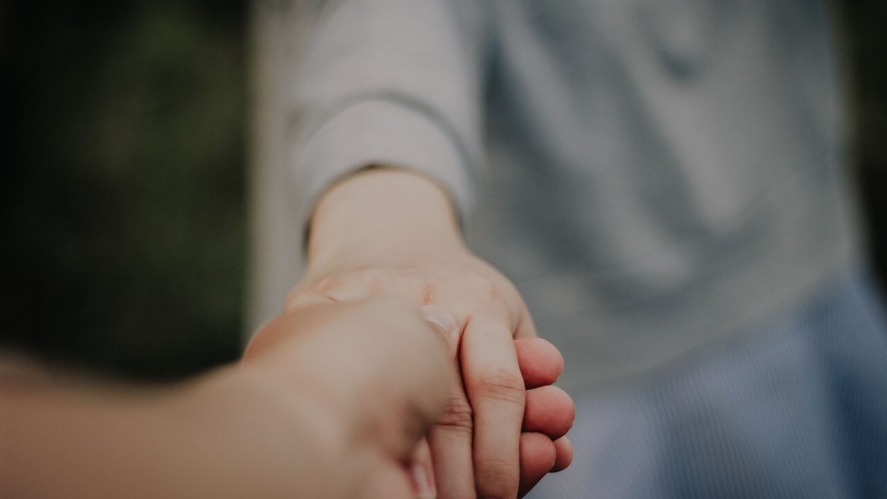 Zwei Hände halten sich fest. Eine Hand greift die andere, symbolisiert Unterstützung und Verbindung zwischen den Personen. Der Hintergrund ist verschwommen, wodurch die Berührung in den Fokus rückt.