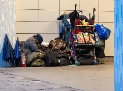 Eine Person sitzt zusammengekauert auf dem Boden, umgeben von Gepäck und Rucksäcken. Ein Einkaufswagen, beladen mit verschiedenen Taschen und persönlichen Gegenständen, steht daneben. Die Szene vermittelt ein Gefühl von Obdachlosigkeit und Entbehrung in einer urbanen Umgebung.