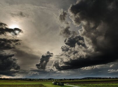 Ein dramatischer Himmel mit dunklen, bedrohlichen Wolken und einem Hauch von Sonnenlicht, der durchbricht. Im Vordergrund führt ein Weg durch eine weite Landschaft, umgeben von Feldern und sanften Hügeln. Die Atmosphäre vermittelt ein Gefühl von Spannung und bevorstehendem Wetterwechsel.
