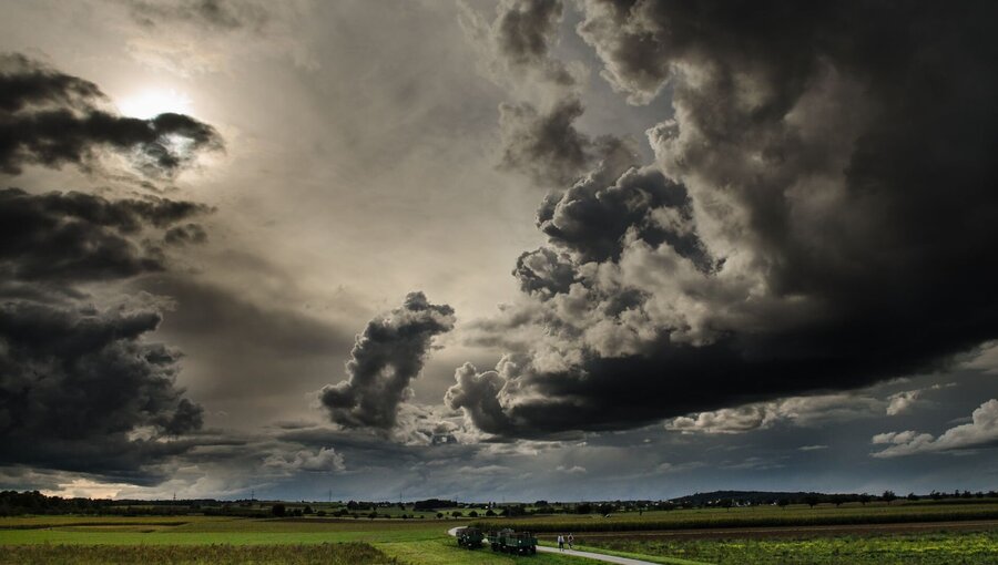 Ein dramatischer Himmel mit dunklen, bedrohlichen Wolken und einem Hauch von Sonnenlicht, der durchbricht. Im Vordergrund führt ein Weg durch eine weite Landschaft, umgeben von Feldern und sanften Hügeln. Die Atmosphäre vermittelt ein Gefühl von Spannung und bevorstehendem Wetterwechsel.