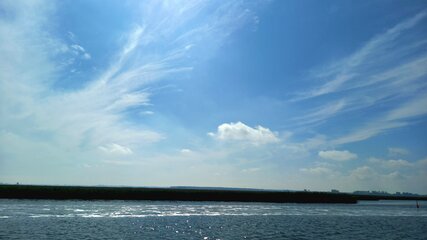 Ein weites Gewässer erstreckt sich im Vordergrund, das von der Sonne beleuchtet wird und glitzert. Der Himmel ist blau mit einigen weißen Wolken, während am Horizont ein dunklerer Streifen, vermutlich ein Ufer oder ein Damm, sichtbar ist.